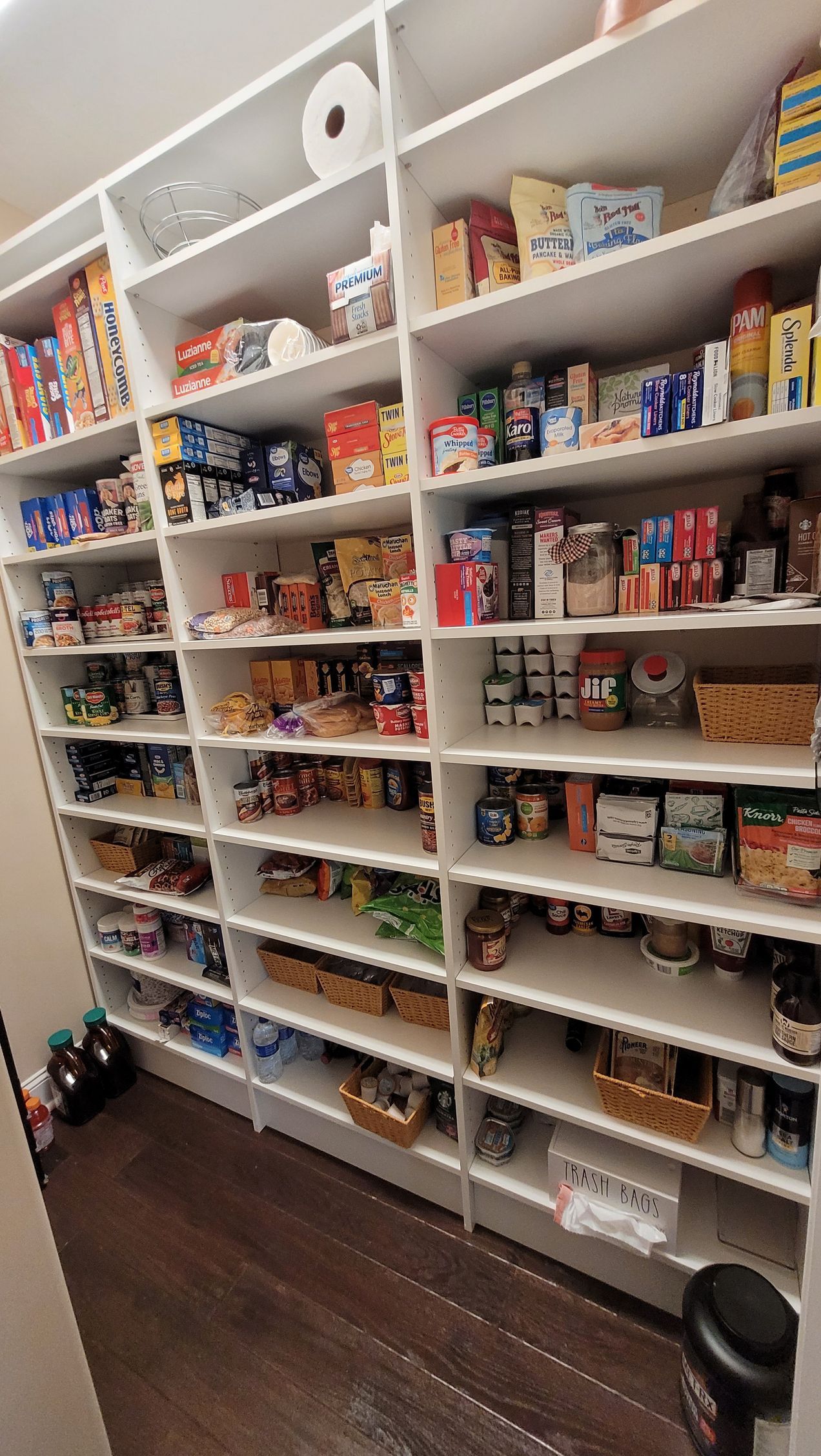 Walk-in pantry with white shelves stocked with food cans, boxes, and jars on a dark wood floor.
