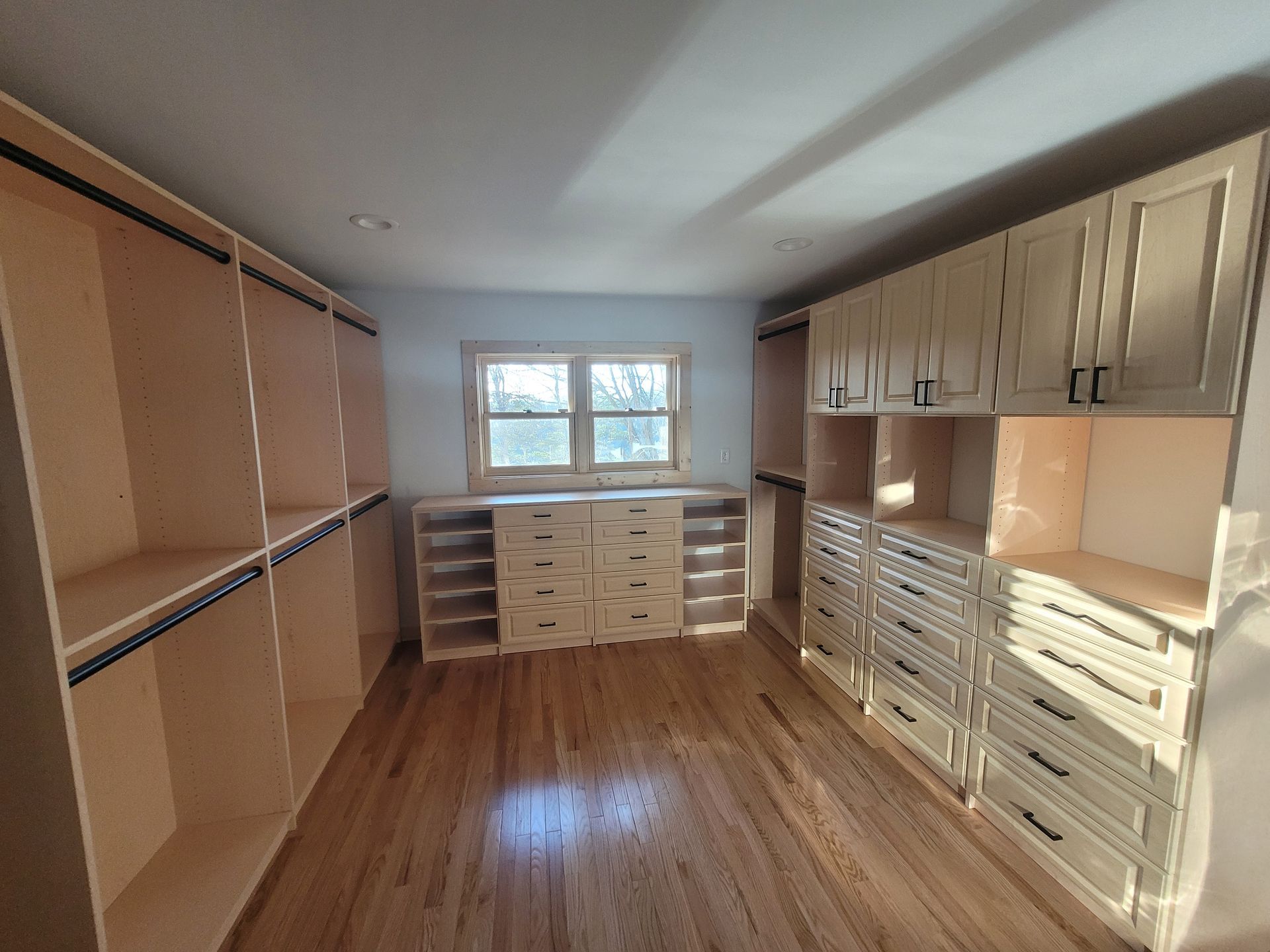 Empty walk-in closet with wood floors, beige cabinets, and drawers beneath a window.