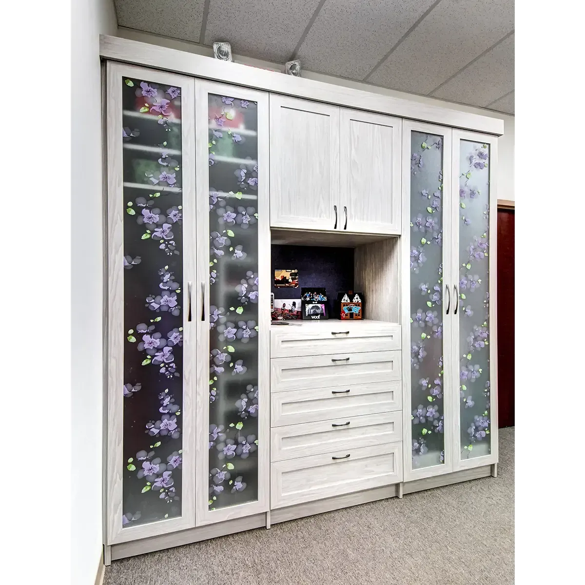 White bedroom wardrobe with floral glass doors, central drawers, and small items displayed on the middle shelf