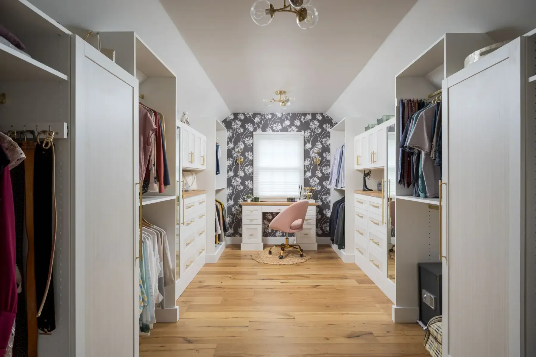 Bright walk-in closet with open white wardrobes, wooden floor, and a desk by a window with floral wallpaper.