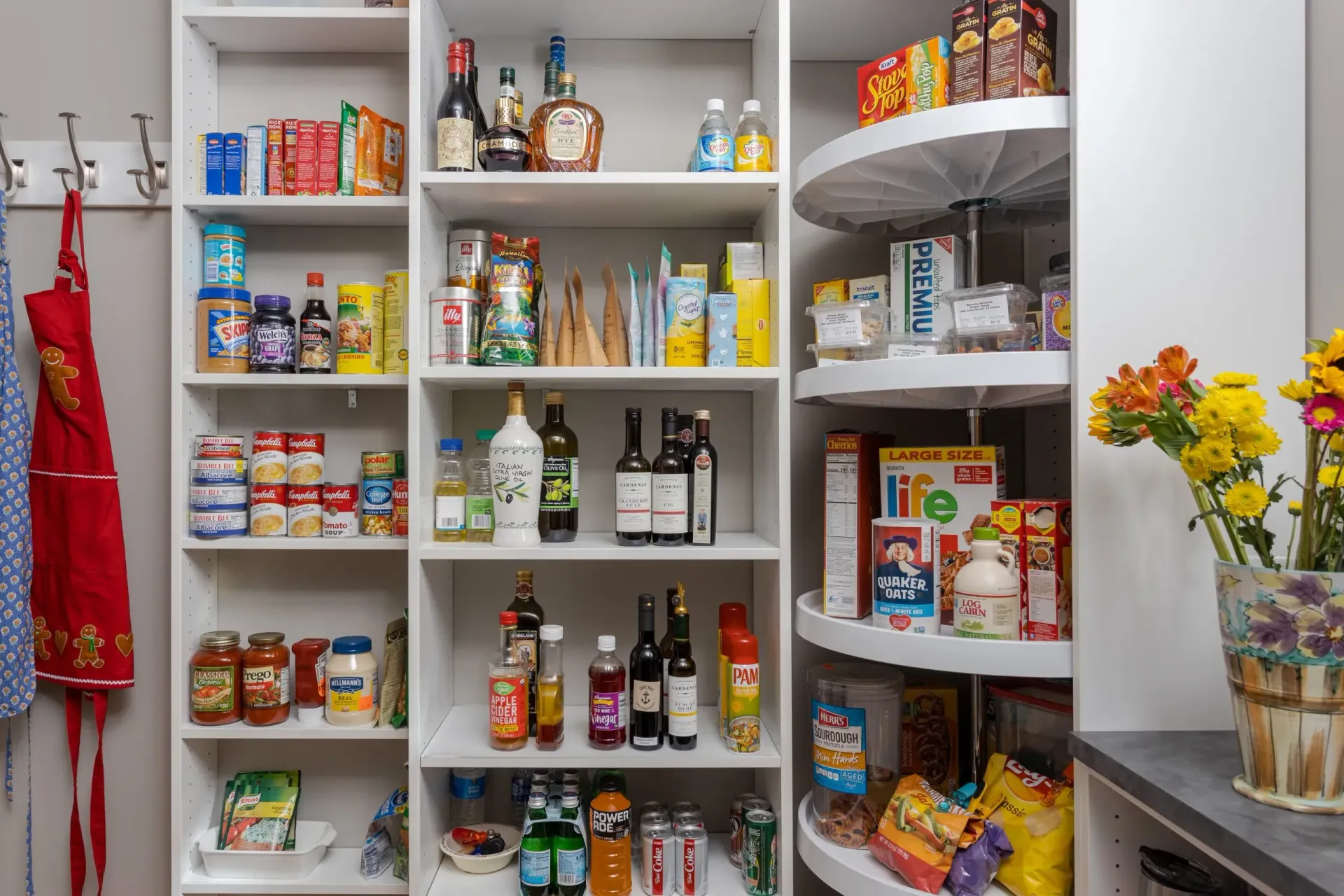 Open pantry shelves stocked with snacks, bottles, and canned goods, with a vase of yellow flowers on the right.