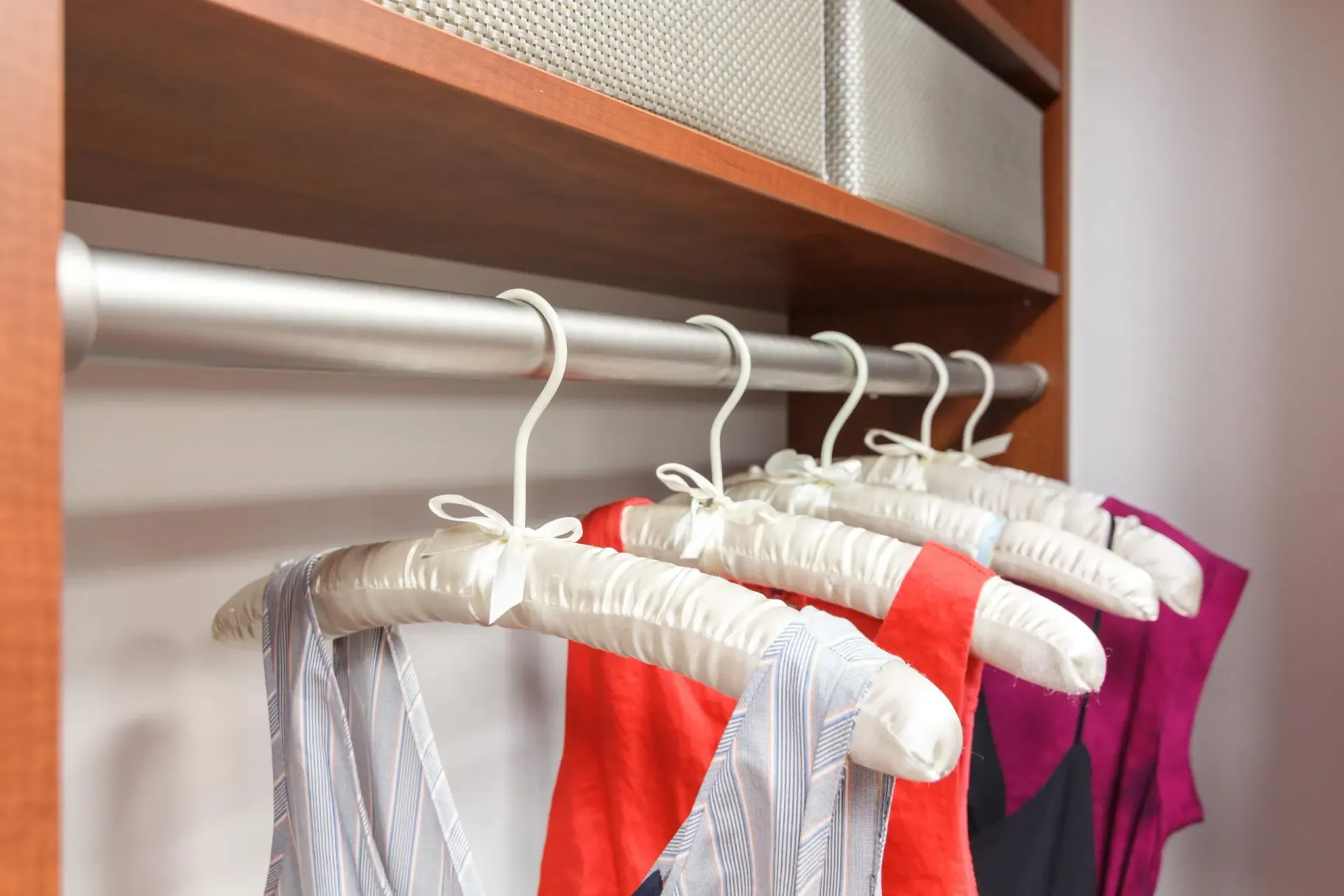 Clothes hang on white hangers in a wooden closet shelf, including silver, coral, white, and magenta garments