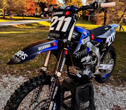Blue dirt bike numbered 211 parked on a gravel path with autumn trees in the background