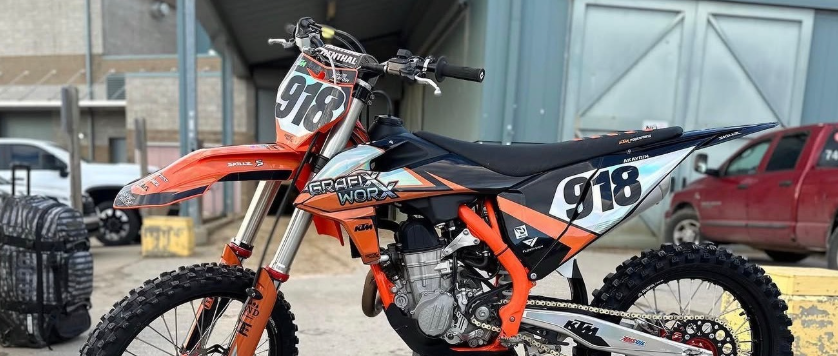 Orange dirt bike with number 918 parked indoors on a stand, with a red car in the background.