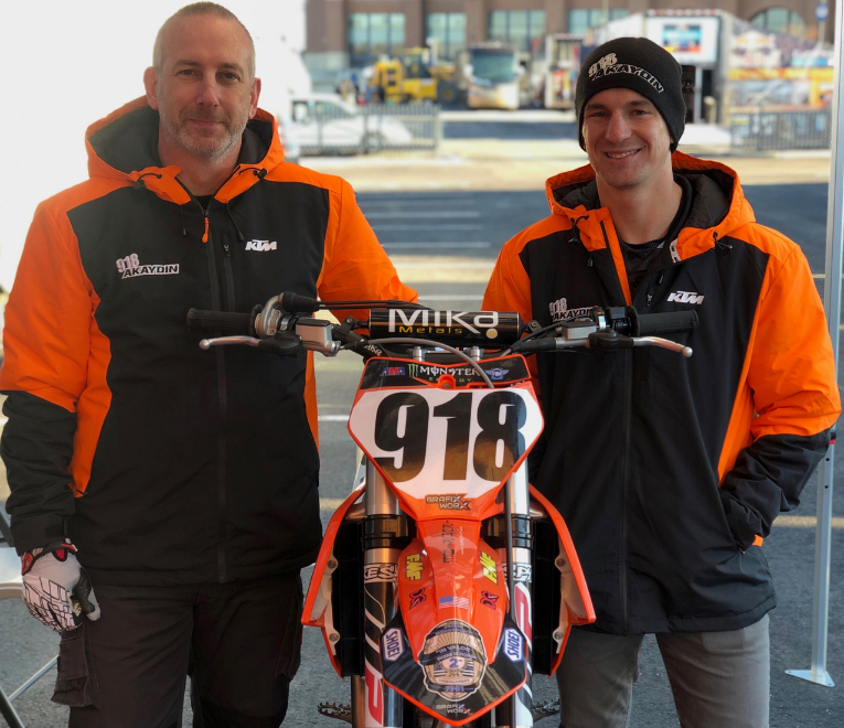 Two people in orange jackets stand behind a red dirt bike numbered 918 indoors.