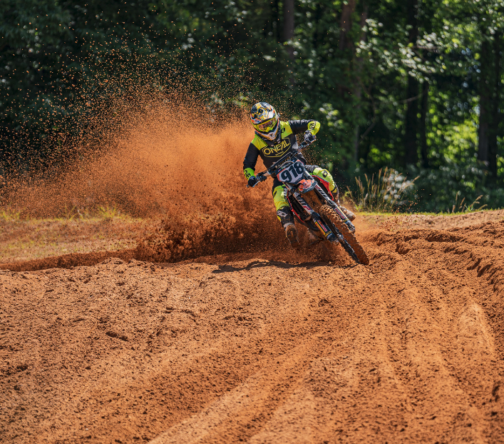 Motocross rider leaning into a dirt track turn, kicking up a cloud of orange dust.