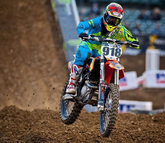 Motocross rider in blue gear jumps over dirt track on red bike, race number 918 visible.