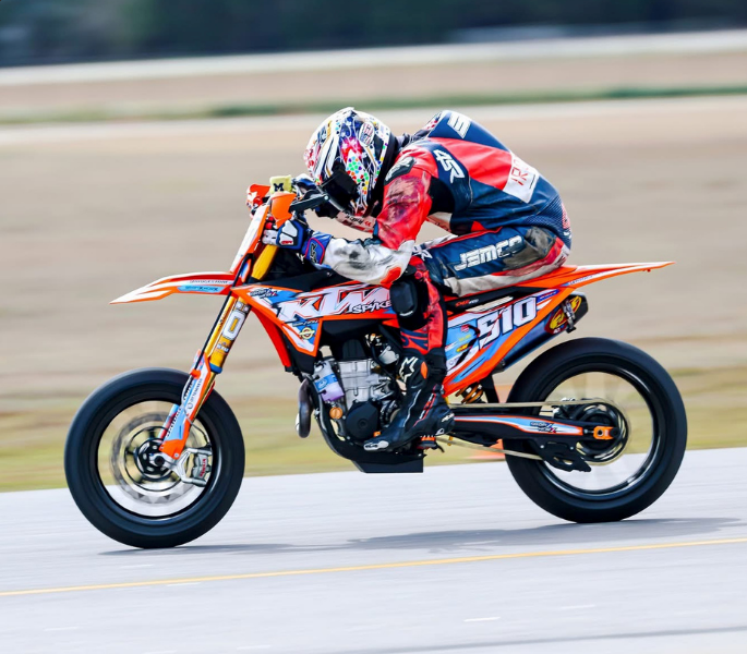 Motorcyclist in red and blue racing gear leans low on an orange sport bike at speed on a road