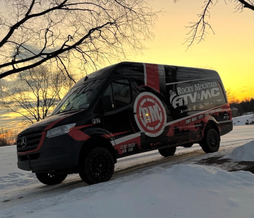Black branded cargo van parked on snowy ground at sunset beneath bare trees.