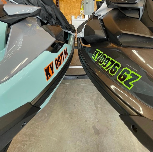 Two jet skis parked side by side, gray with orange lettering and brown with green lettering, in a garage