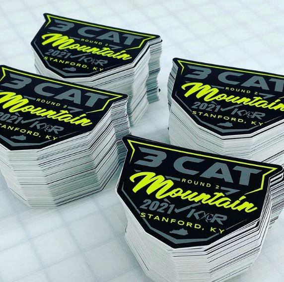 Stacks of black, yellow, and white 3CAT Mountain sticker labels on a light surface.