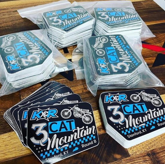 Stacks of “3 Cat Mountain” stickers in blue, black, and white on a wooden table.