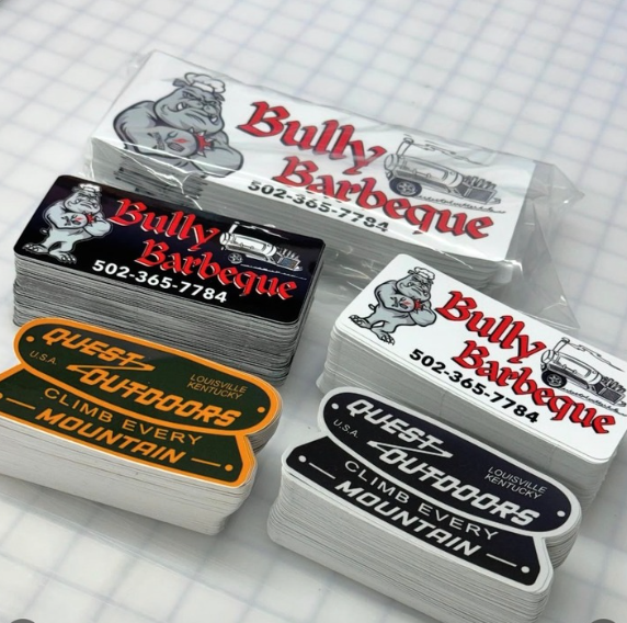 Stacked custom bumper stickers for Bully Barbeque and Quest Outdoors on a gridded surface