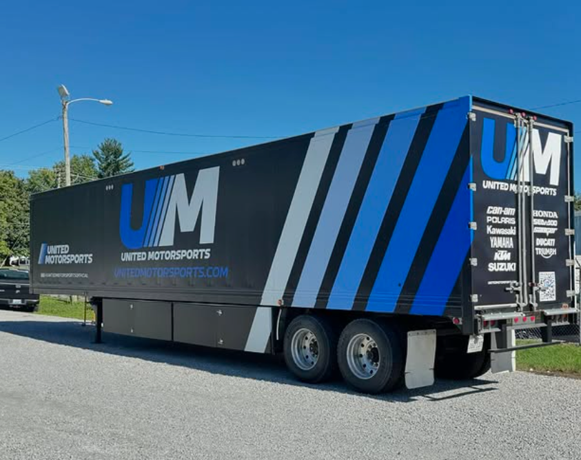 Black UM utility trailer with blue stripes parked outdoors on a sunny day