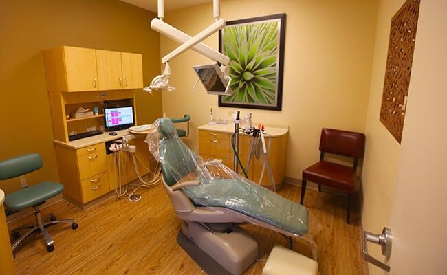 Dental exam room with reclining chair, overhead light, cabinets, computer, and framed wall art