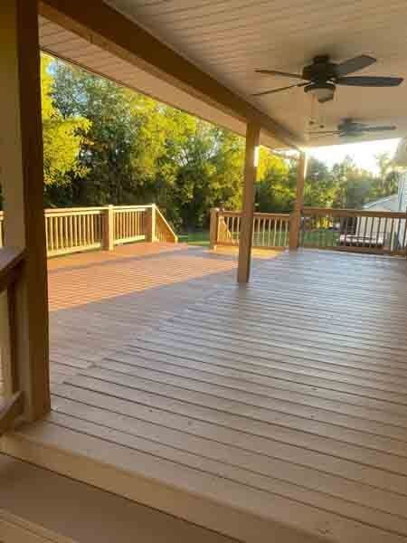 Covered wooden deck with railing, ceiling fan, and trees in the background