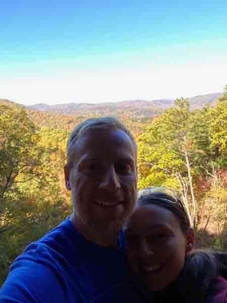 Two people smiling in front of a bright autumn forest and distant mountains