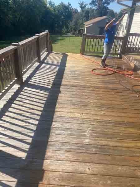 Person pressure-washing a sunlit wooden deck with a hose and orange extension cord