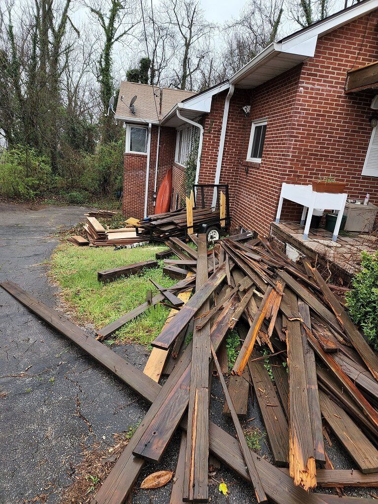 Damaged brick house with piles of broken wooden boards along a wet driveway in a wooded area