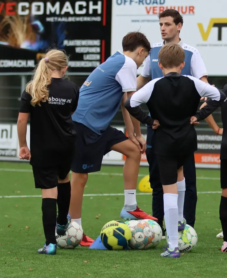 Een voetbalcoach en jeugdspelers staan bij elkaar rond ballen op een trainingsveld.