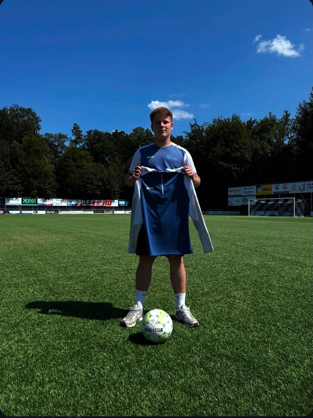 Jongere op een voetbalveld met een blauw voetbalshirt in de hand en een voetbal aan de voeten, onder een strakblauwe hemel