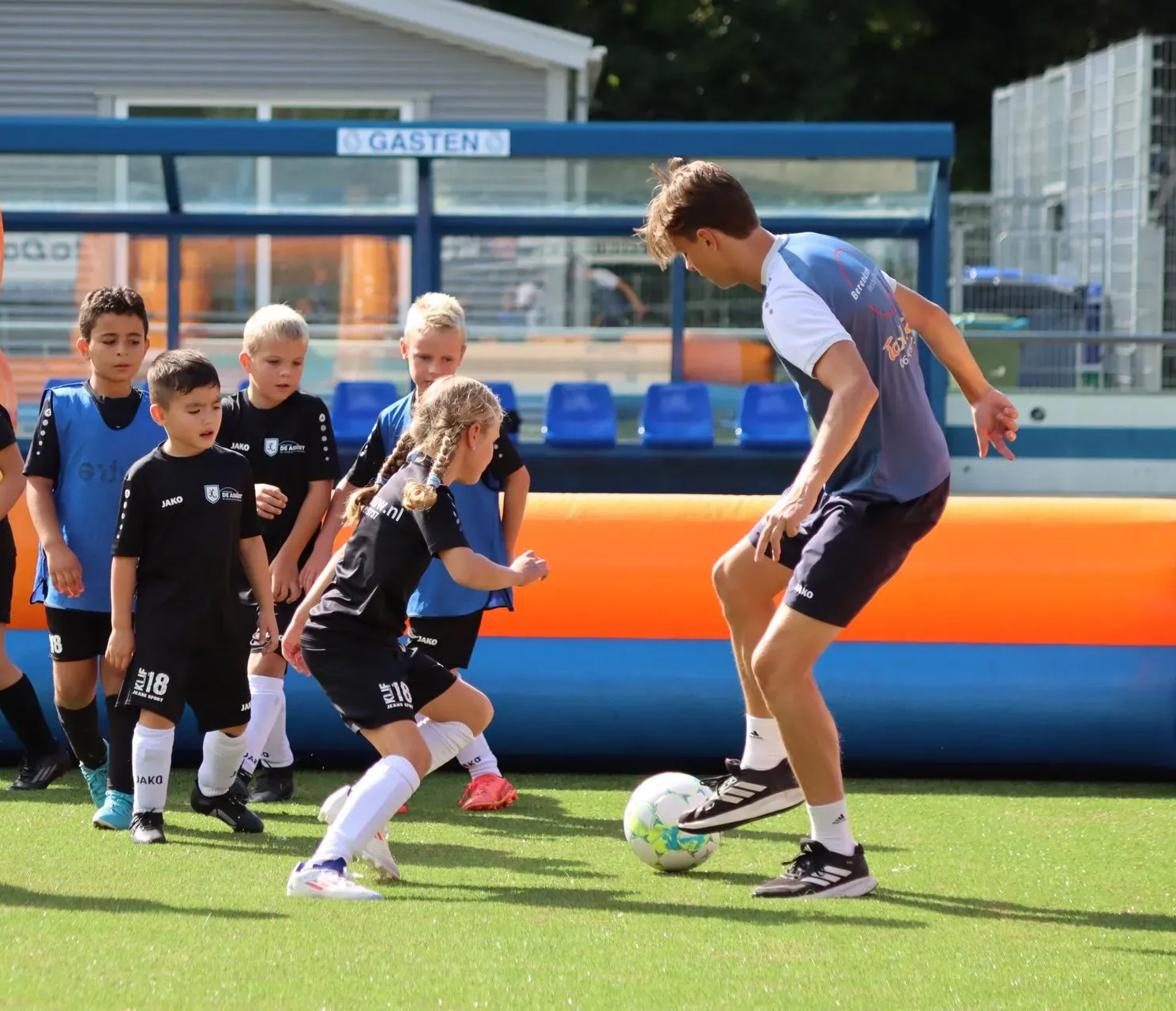 Kinderen spelen voetbal op een veld, terwijl een coach naast hen een bal dribbelt en er op de achtergrond blauw-oranje bankjes staan.