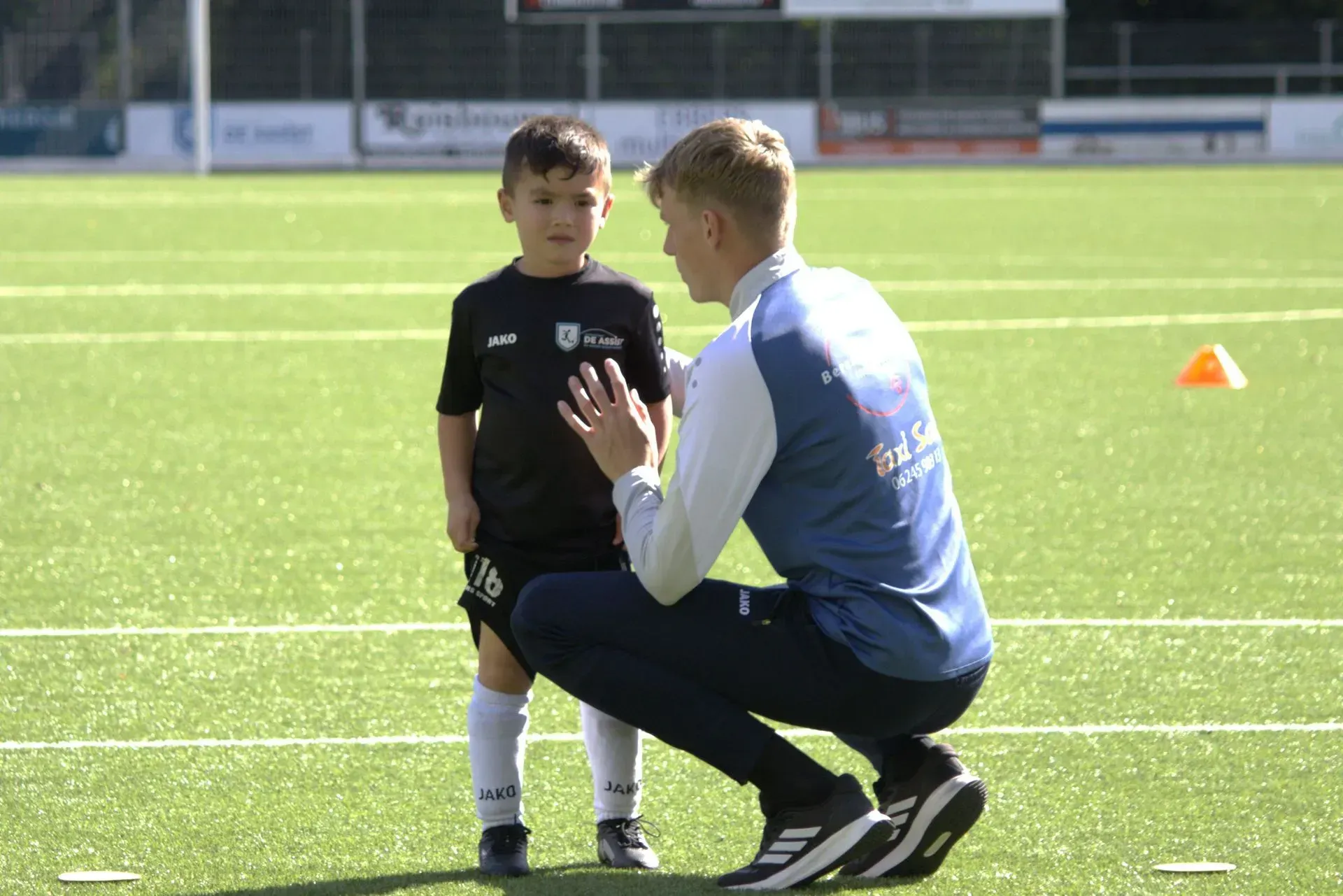 Een coach knielt naast een kind op een voetbalveld en praat met hem tijdens de training.
