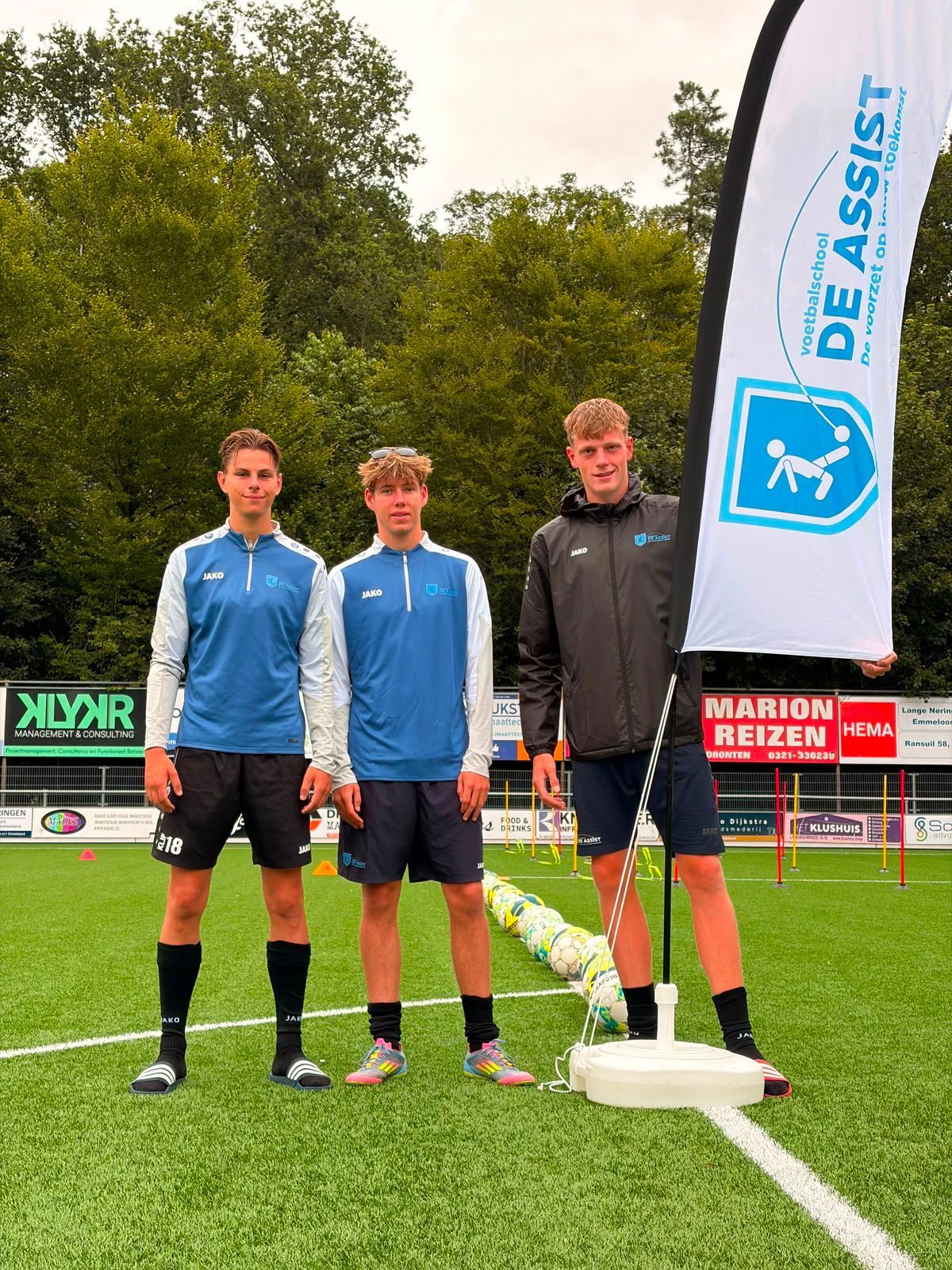 Twee sporters in blauwe shirts staan op een voetbalveld naast een DE ASSIST-vlag.