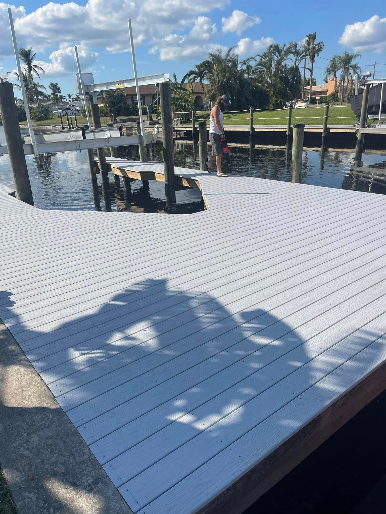 White dock by calm water with a person standing near the edge, boats and palm trees in the background.