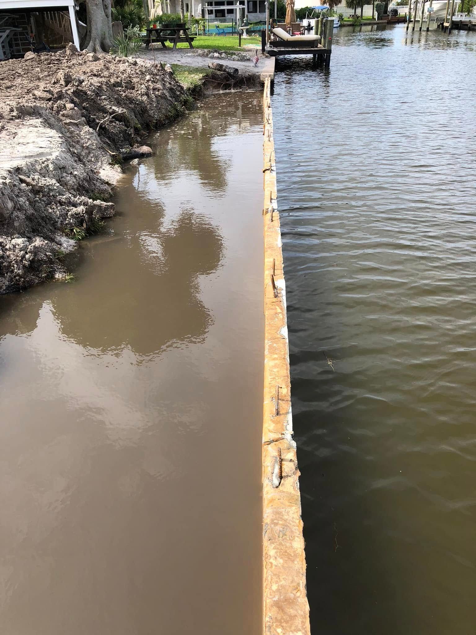 Construction barrier dividing muddy water and clear water in a canal, with a small dock and houses in the background