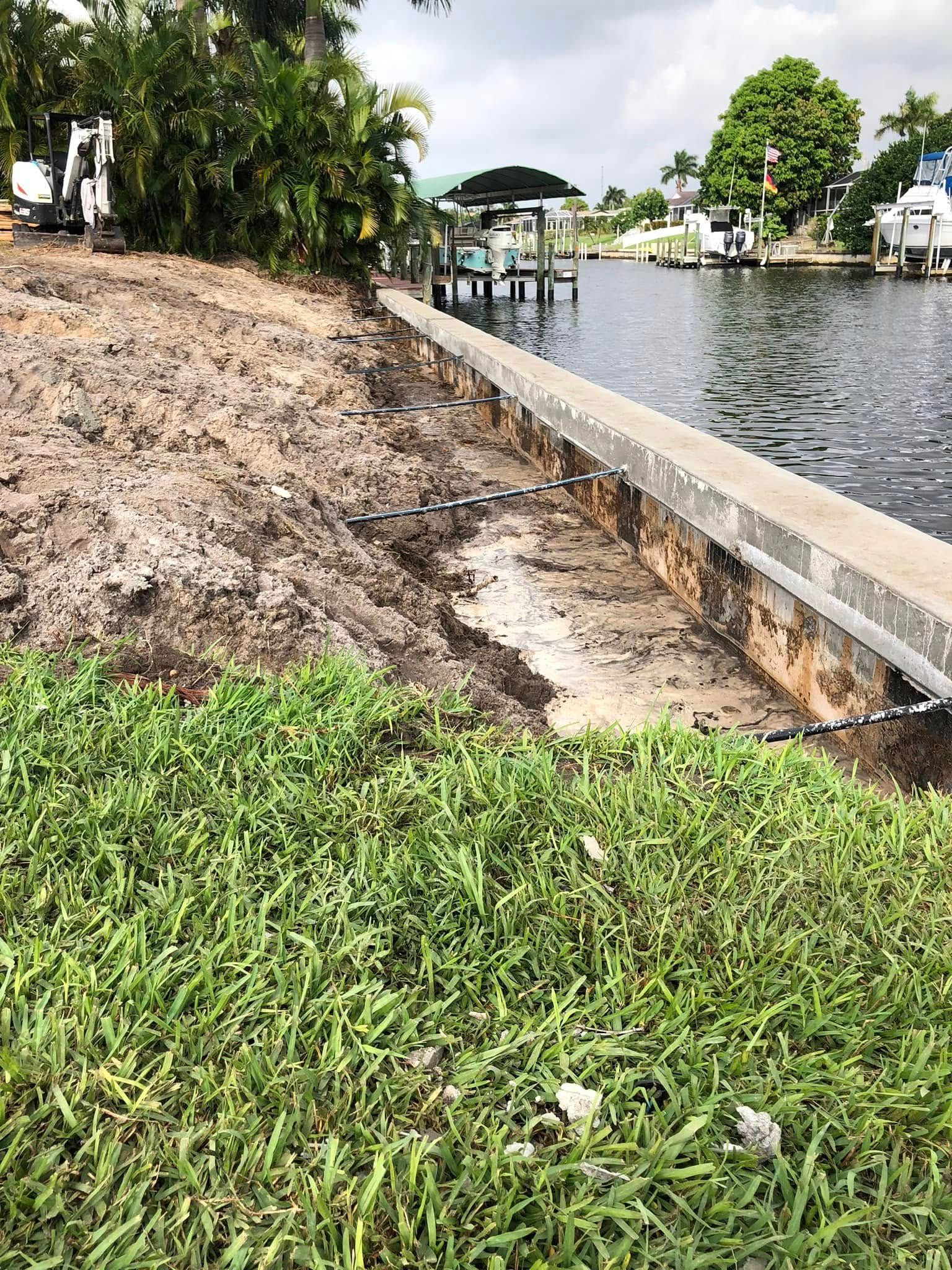 Eroded grassy riverbank beside a canal, with a concrete edge and small dock in the distance.