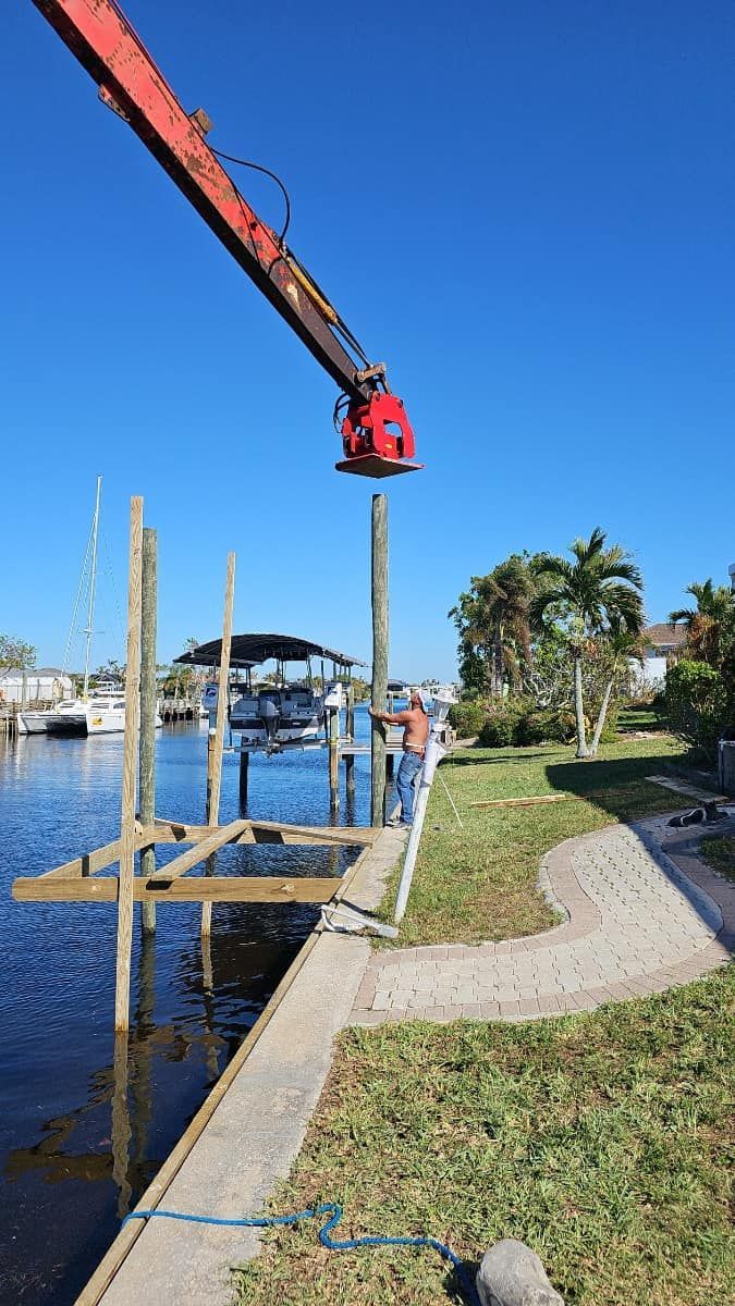 Crane lifting a dock frame beside a canal, with grassy shoreline and palm trees under a clear blue sky