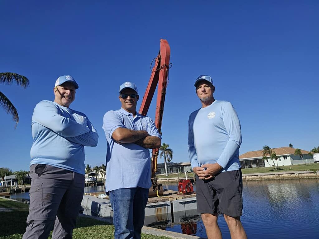 Three people in light blue shirts posing by a waterfront under a clear blue sky.