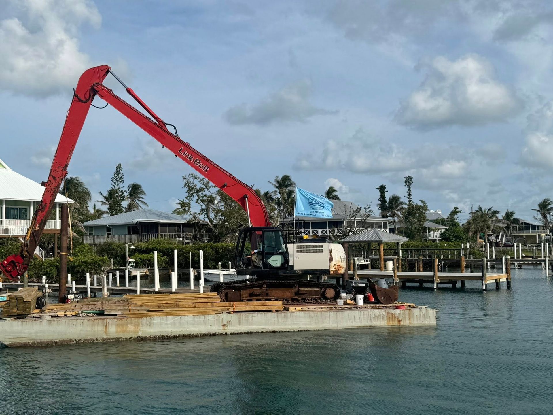 Red excavator on a barge by the waterfront, lifting debris near a dock under a cloudy sky