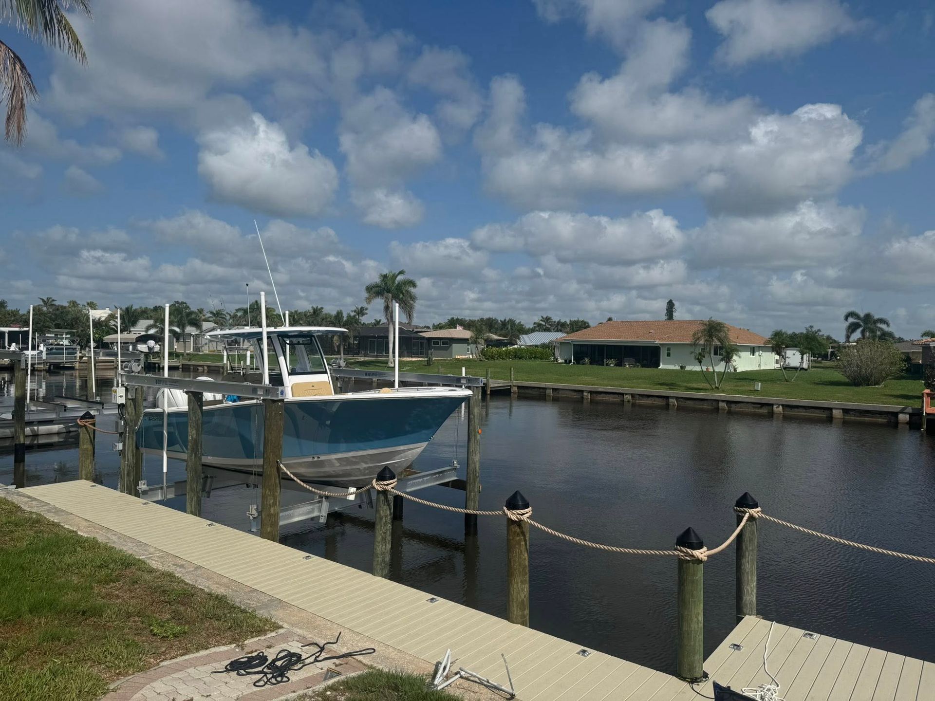 Boat dock on a calm canal with ropes, palm trees, and a house under a cloudy blue sky