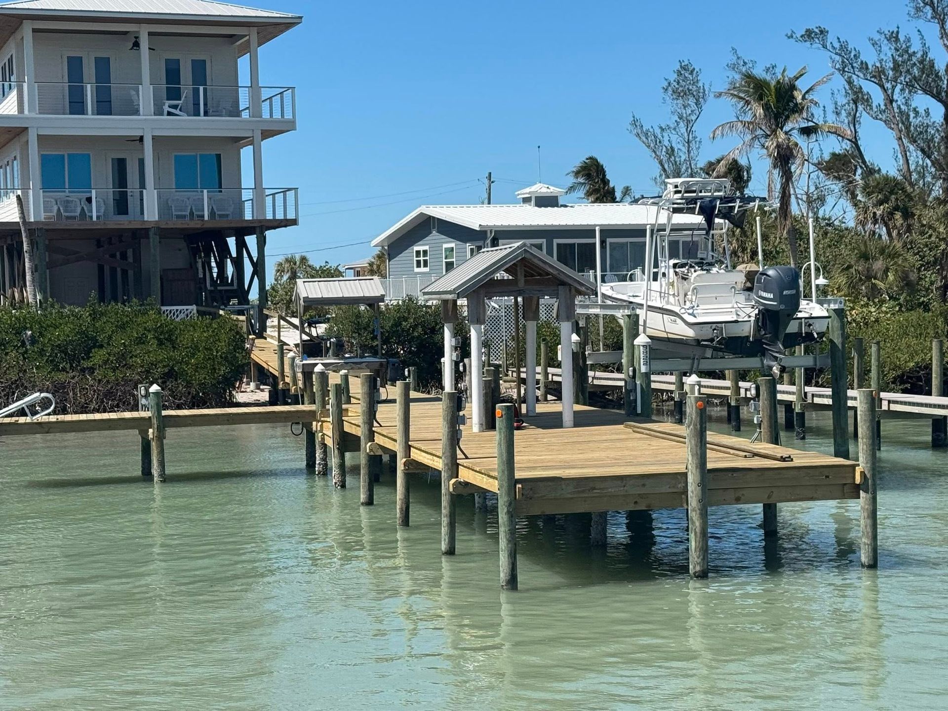 Waterfront dock with wooden pilings, clear green water, and coastal houses under a blue sky