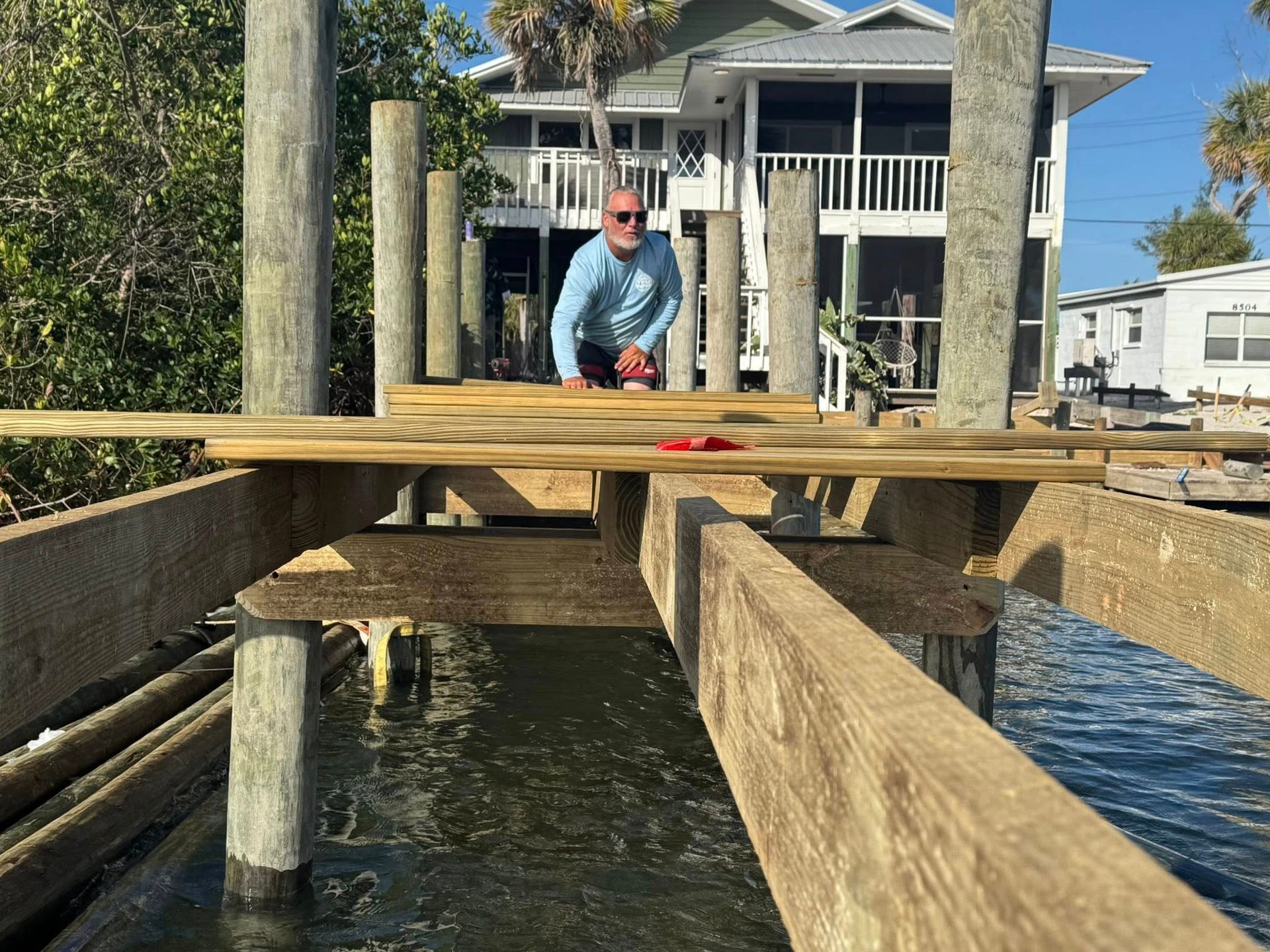 Man in blue shirt working on wooden dock pilings beside a canal, with a waterfront house in the background