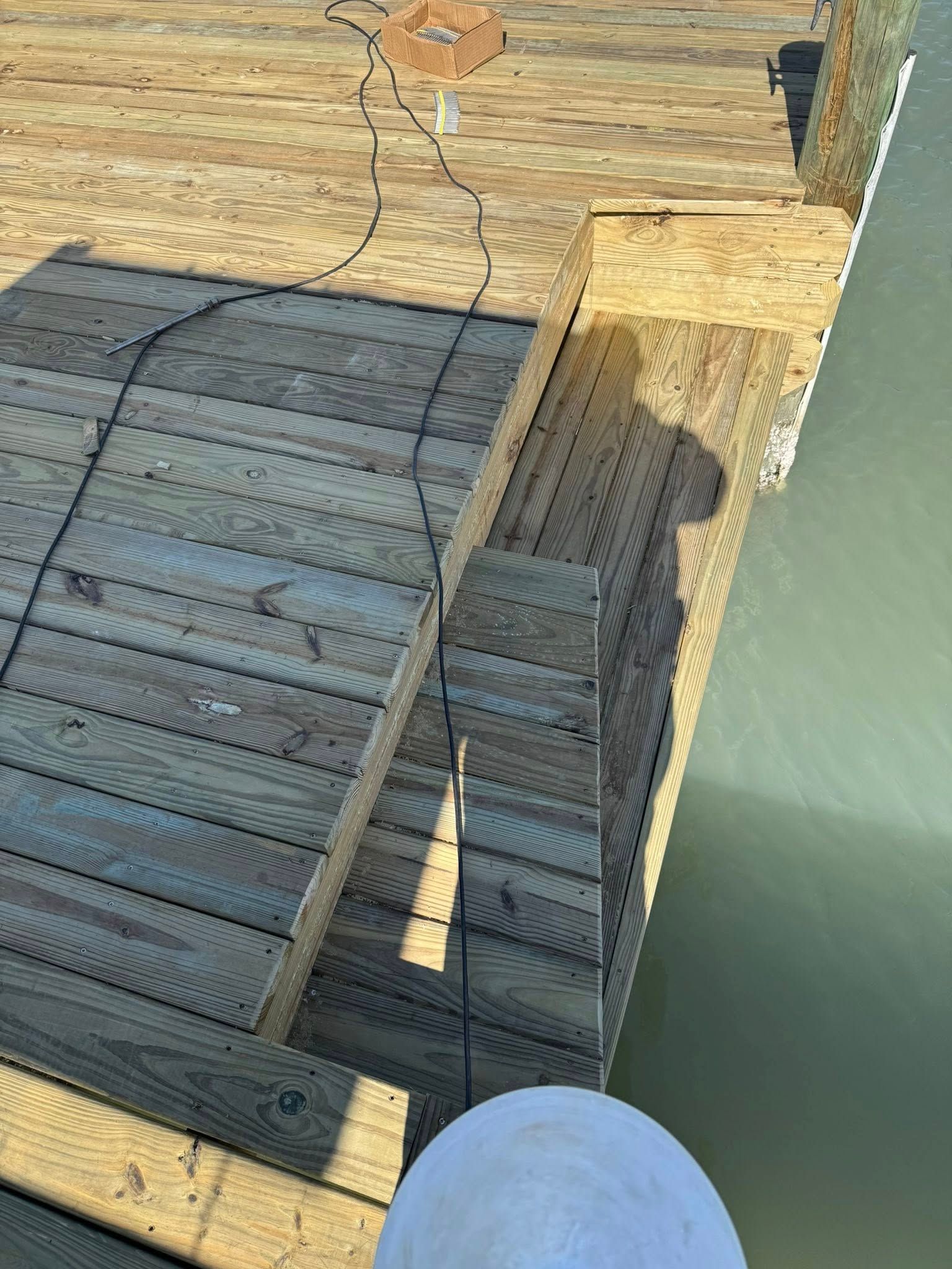 Worn metal ramp on a wooden dock beside green water, with a person’s shadow cast across it