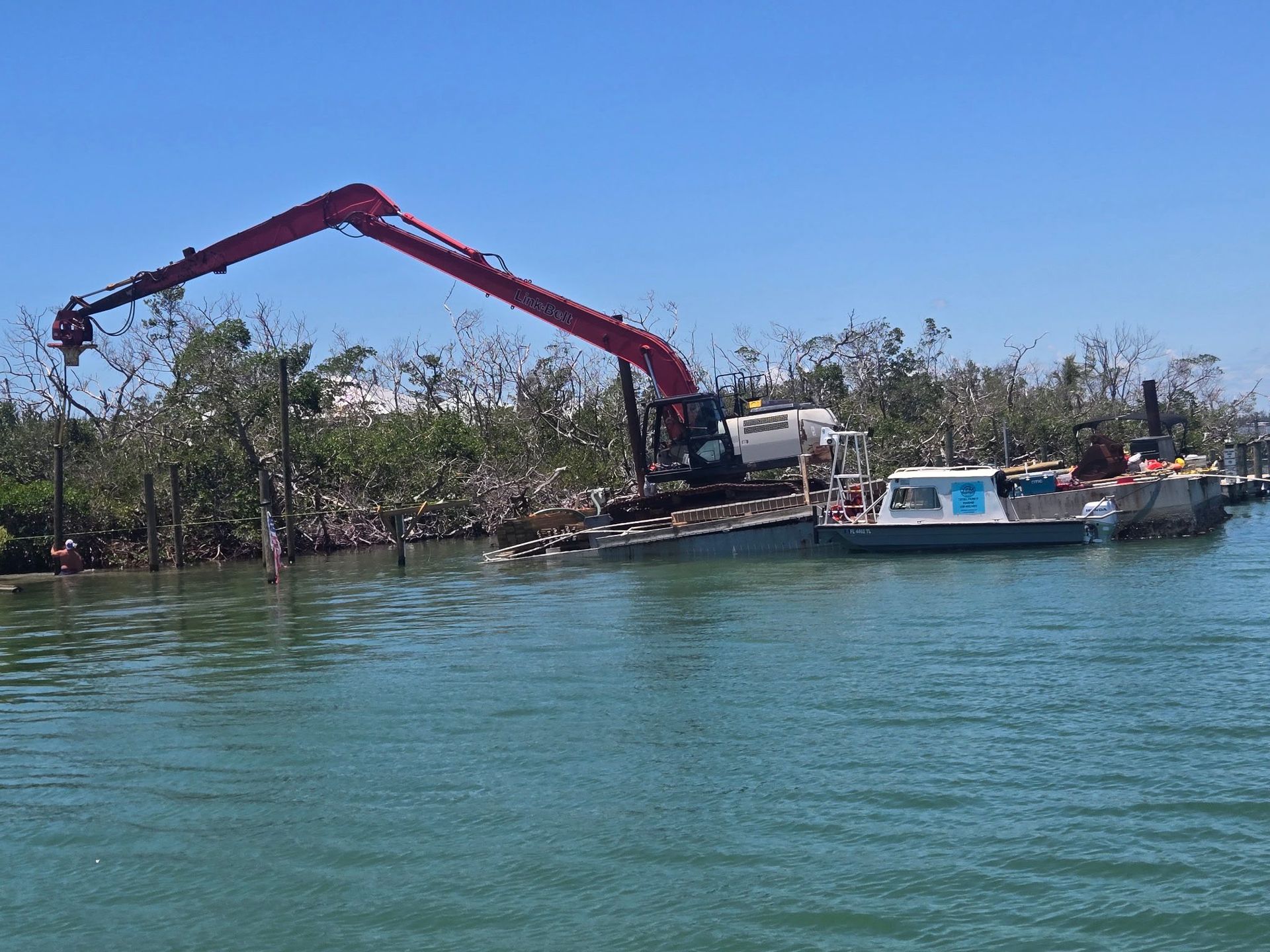 Red crane barge working beside a small boat on calm blue water near a mangrove shoreline