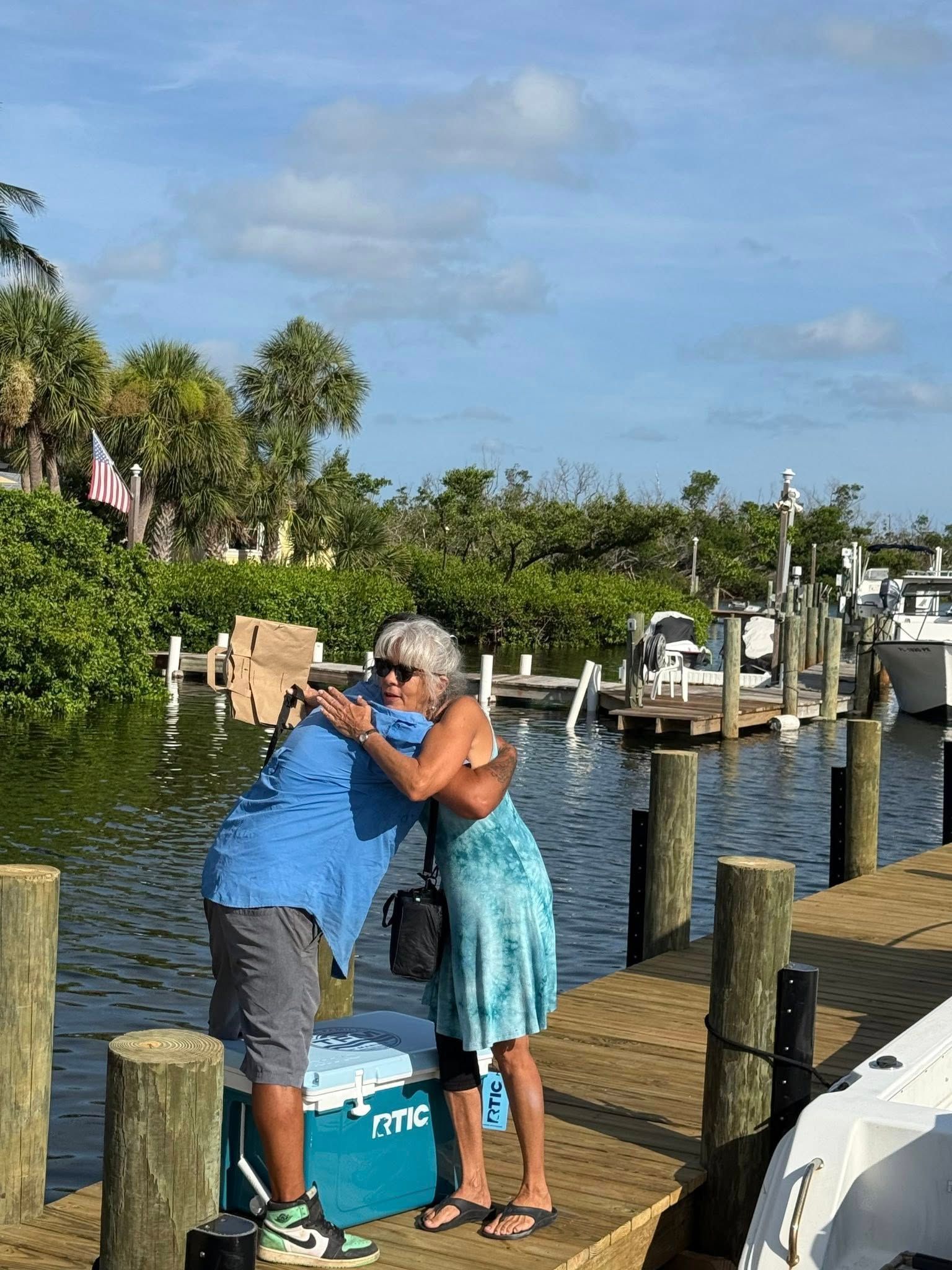 Two people hugging on a dock by the water, with boats and pilings in the background.