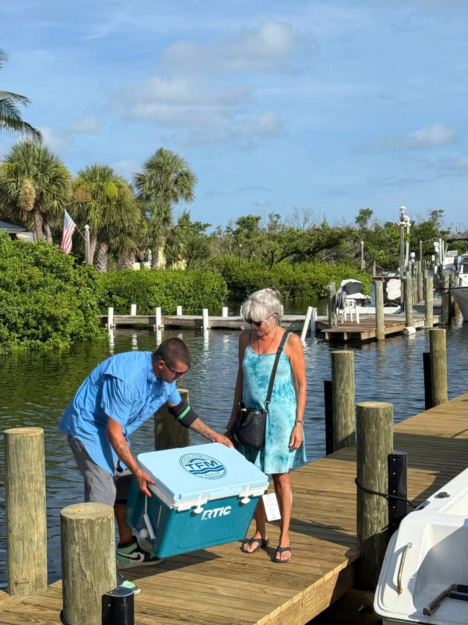 Man unloading a blue cooler on a wooden dock beside a woman near a waterfront with boats and palms