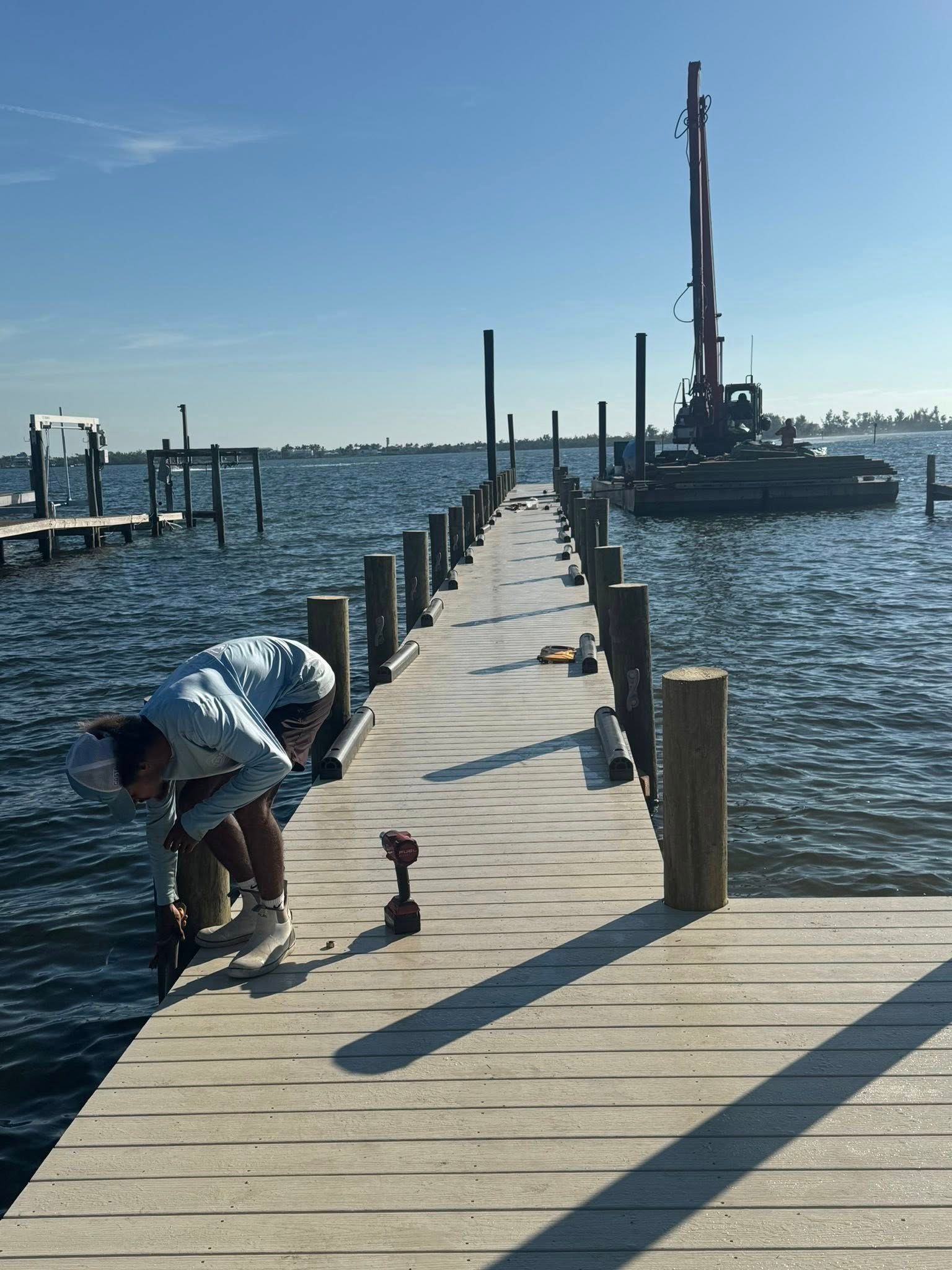 Person crouches on a wooden dock by the water, with boats and marina equipment in the distance.