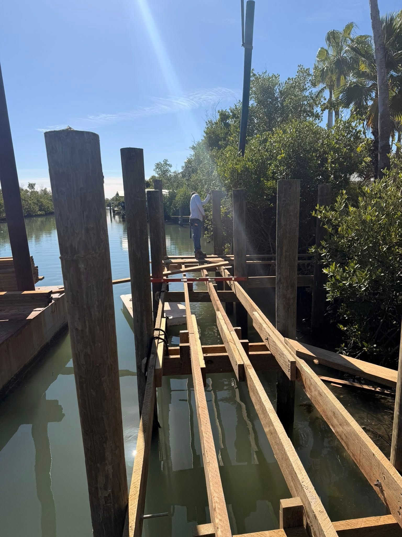 Narrow wooden dock with rails over calm water, viewed under bright blue sky and trees.
