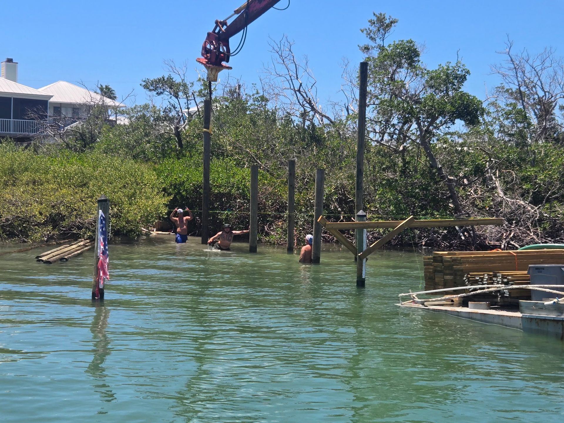 People on a dock in green water near mangroves, with a crane overhead and small boats nearby