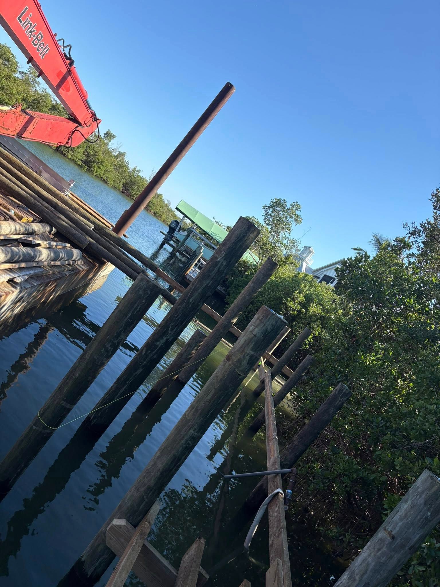 Tilted view of a waterfront dock with wooden posts, trees, and a red crane by calm water under a blue sky