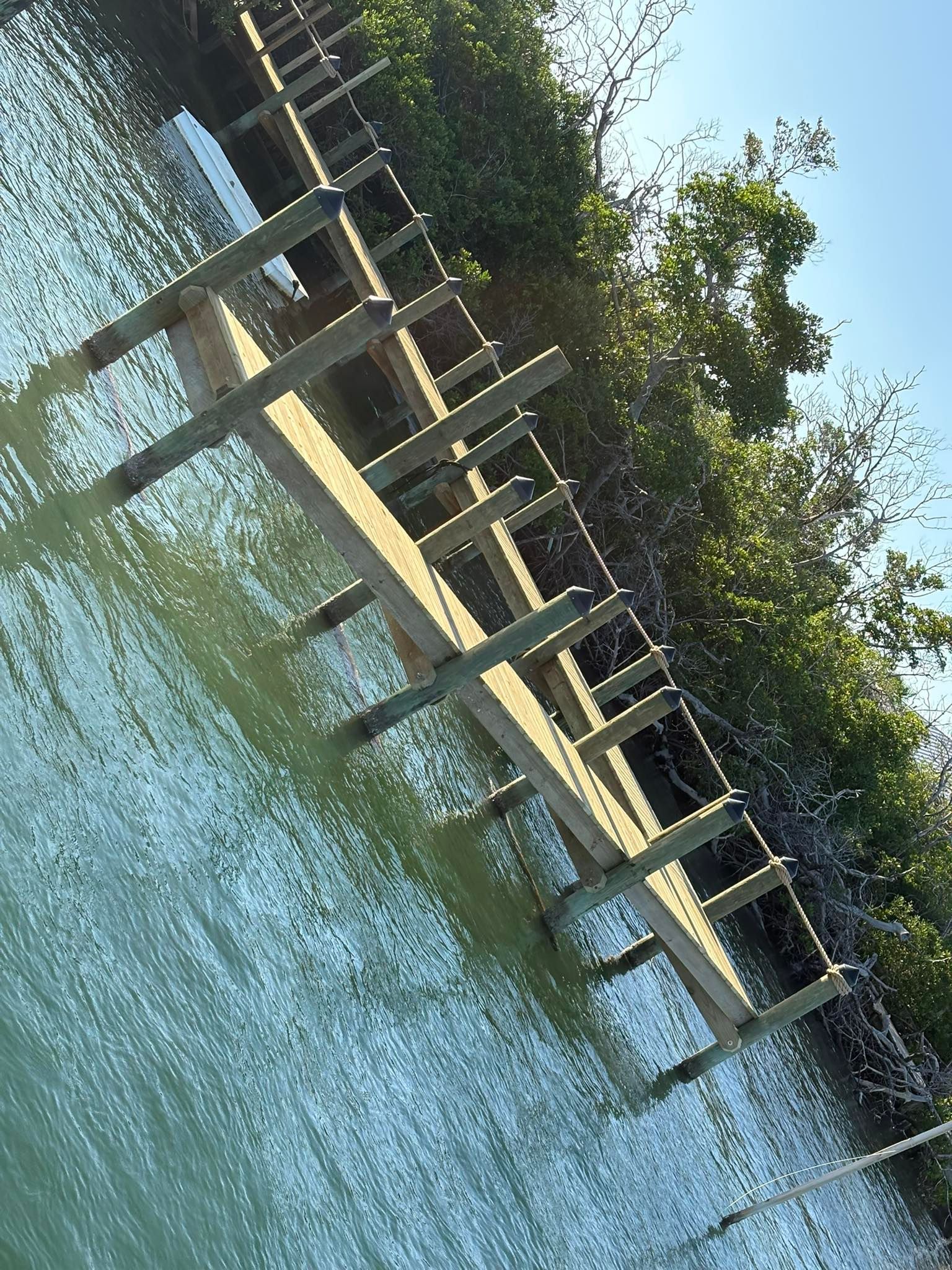 Wooden dock extending over calm blue water beside green trees, reflected in the water.
