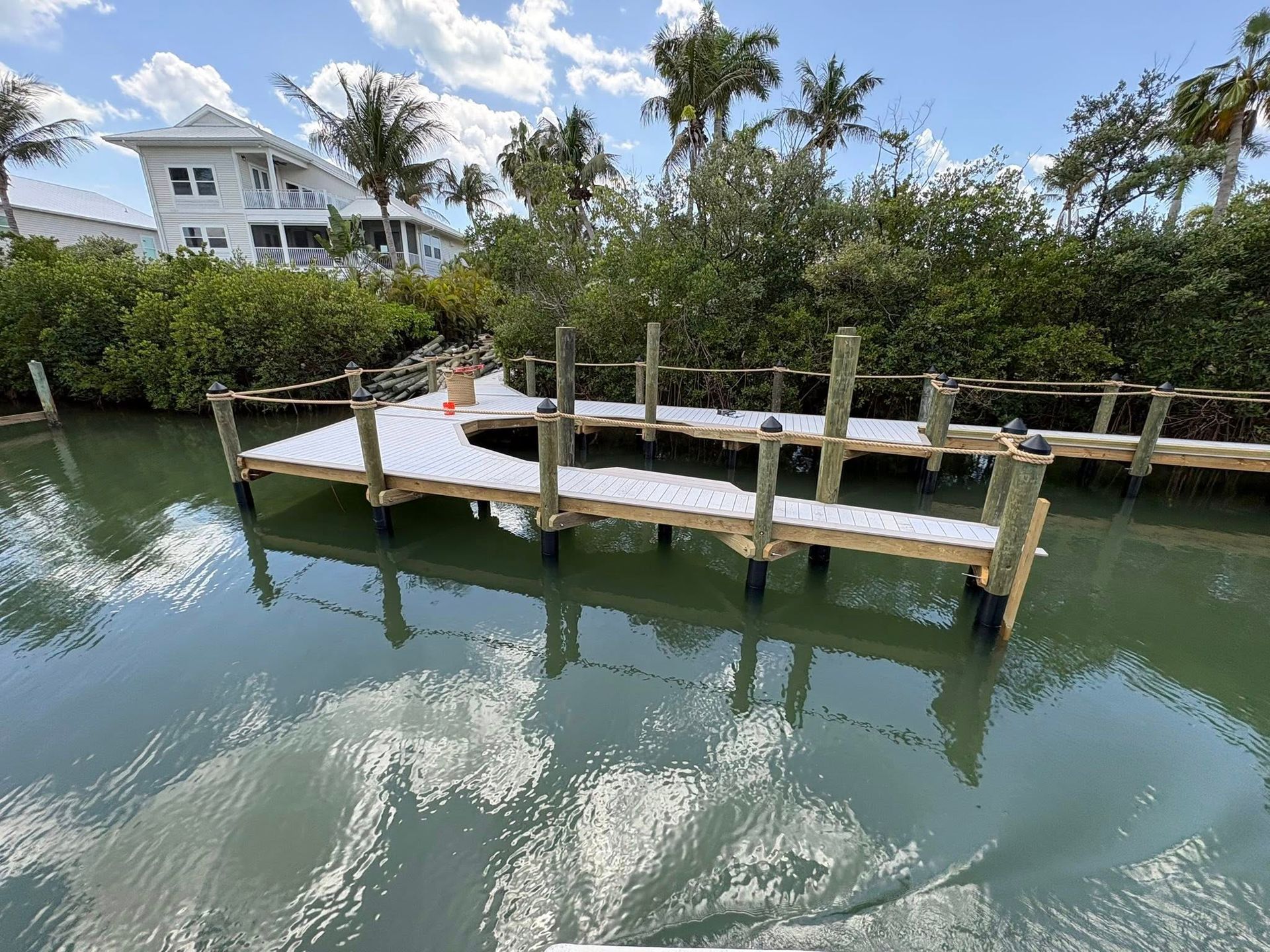 Wooden dock over calm green water beside lush trees and white buildings under a cloudy sky