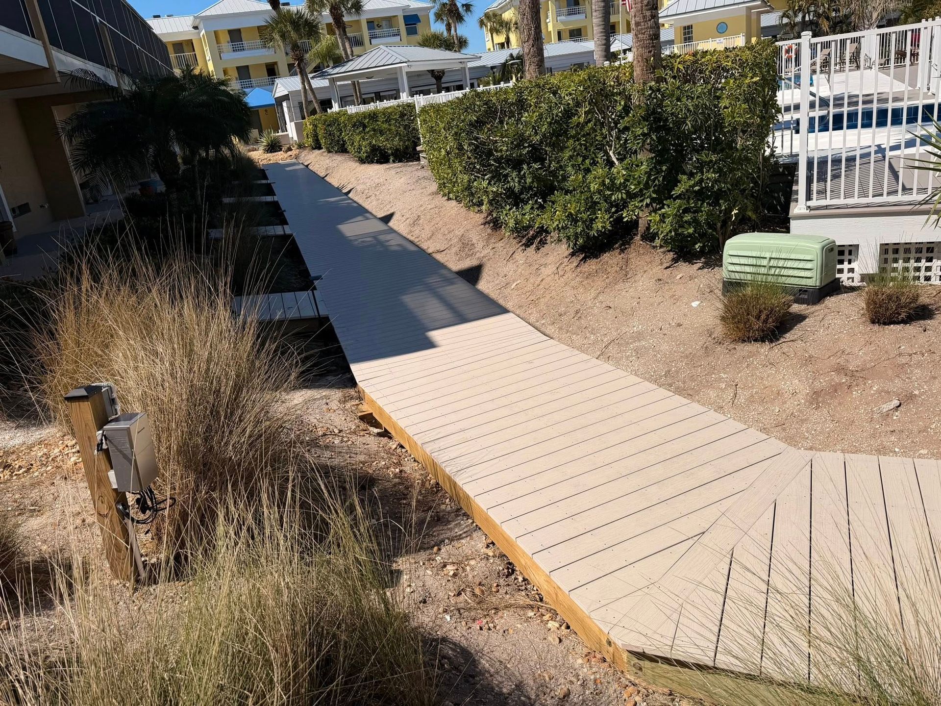 Wooden boardwalk path through desert landscaping beside a fence and shrubs, with buildings in the background