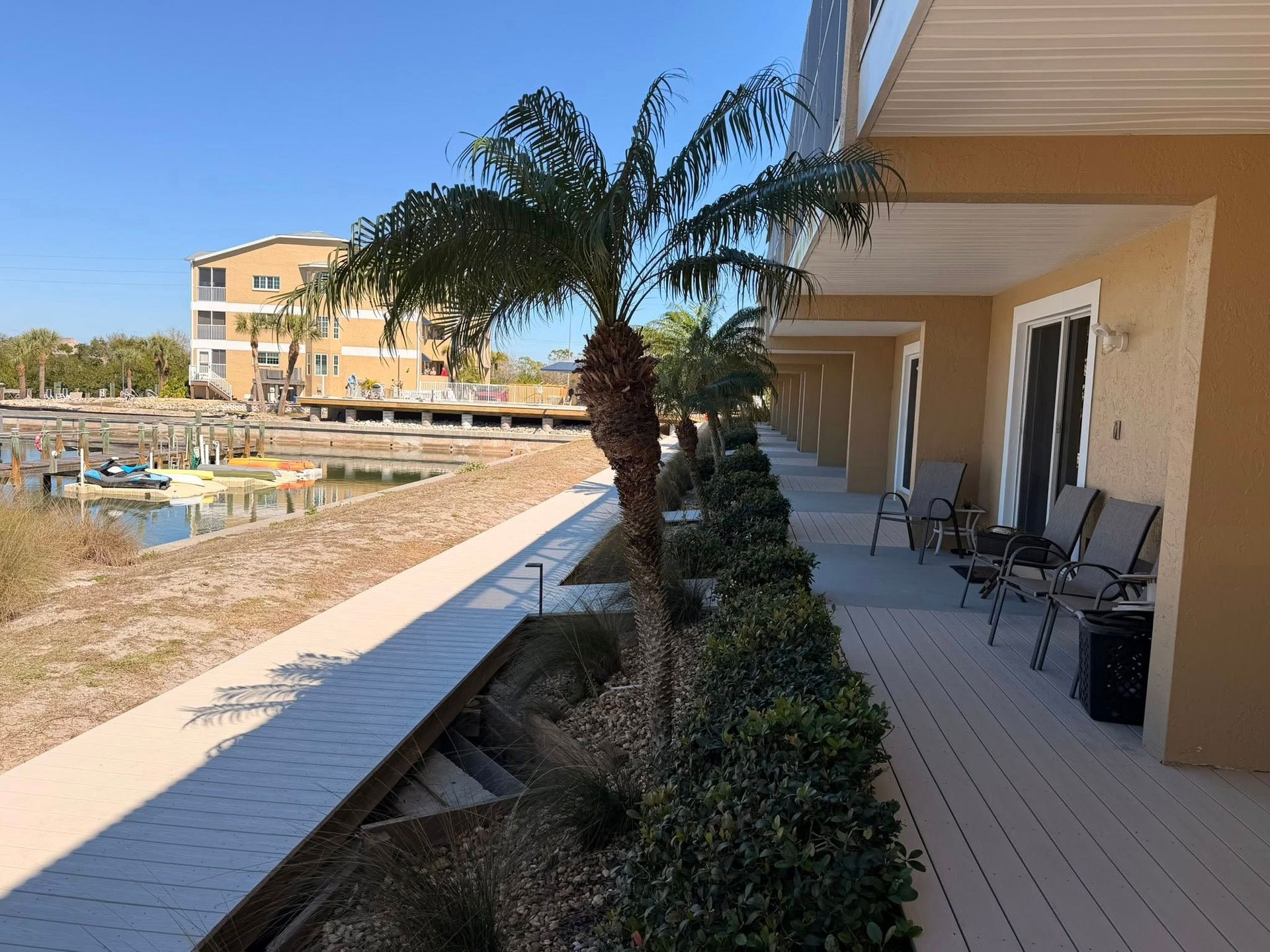 Covered seaside walkway with palm trees, white deck, and chairs beside a waterfront canal