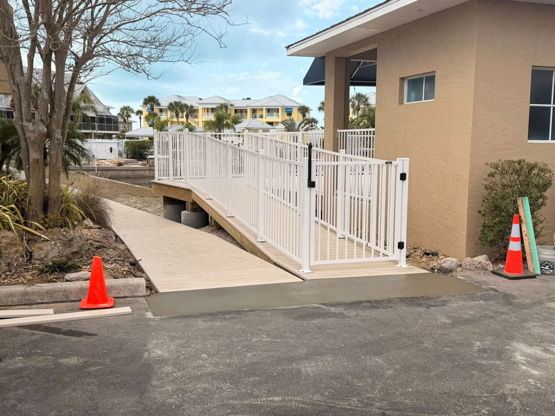 White-railed wheelchair ramp beside a tan building, marked by orange safety cones, with a sidewalk and parking lot.